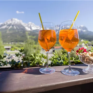 Zwei Gläser mit einem orangefarbenen Getränk stehen auf einem Tisch mit Blick auf schneebedeckte Berge. Neben den Gläsern befindet sich eine Schale mit Snacks.
