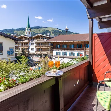 Ein schöner Balkon mit Blick auf eine charmante Stadt. Zwei Getränke und Snacks stehen auf einem Tisch, während die Berge im Hintergrund sichtbar sind.