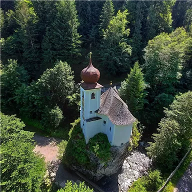 Eine malerische Kapelle auf einem kleinen Felsen, umgeben von dichten Wäldern. Die Kuppel hat eine auffällige rote Farbe und das Gebäude strahlt Ruhe aus.