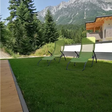 A cozy meadow with two lounge chairs and dense greenery. In the background, majestic mountains rise under a blue sky.
