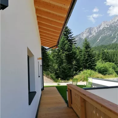 A modern balcony with a wooden terrace and a view of green trees and mountains. The sky is clear and blue.