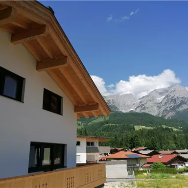 A modern house with wooden cladding and a large balcony. In the background, impressive mountain peaks and green forests can be seen.