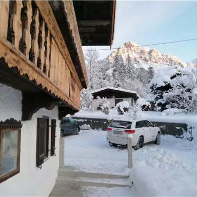 A winter landscape with snow-covered trees and mountains in the background. A traditional wooden house is visible in the foreground, next to a car parked in the snow.