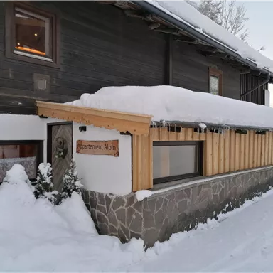 A cozy wooden house with a snow-covered roof in a wintry landscape. The entrance area is simply and invitingly designed.