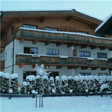 A snow-covered house with a wooden balcony. The garden is surrounded by snow-covered bushes.
