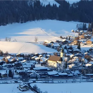A picturesque winter landscape with a small village, framed by snow-covered fields and mountains. At the center stands a church, surrounded by numerous houses.