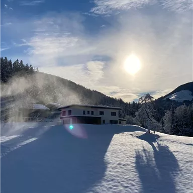 Eine malerische Winterlandschaft mit schneebedecktem Boden und einem modernen Haus. Die Sonne scheint hell am klaren Himmel zwischen sanften Hügeln.