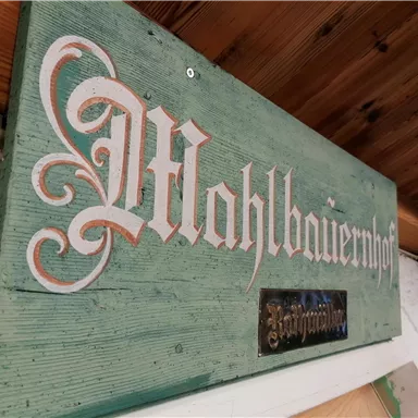 Ein handgemaltes Schild mit der Aufschrift „Mahlbauernhof“ auf grünem Holz. Es hat eine rustikale Ausstrahlung und ist an einer Wand angebracht.