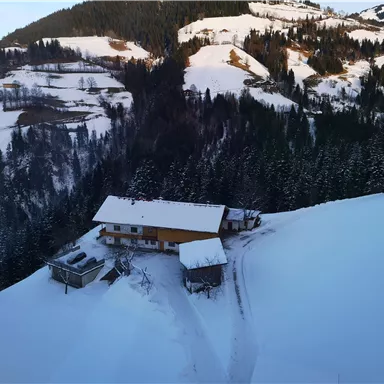 Eine verschneite Landschaft mit einem Haus und Bäumen im Hintergrund. Die Umgebung ist ruhig und idyllisch, umgeben von Bergen.