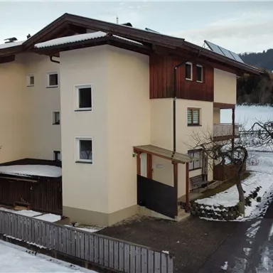 A modern house with wooden elements, surrounded by a snowy landscape. Mountains are visible in the background.