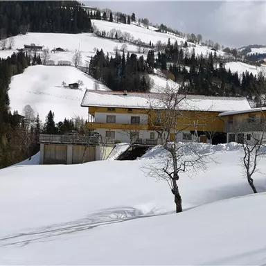 Eine verschneite Landschaft mit einem Haus im Vordergrund. Im Hintergrund sind schneebedeckte Hügel und Bäume sichtbar.