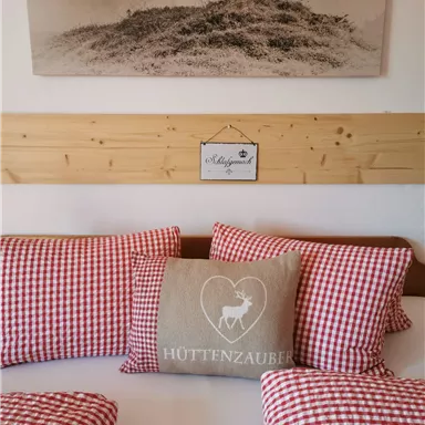 Ein gemütliches Bett mit karierten roten Kissen und einem dekorativen Kissen mit einem Hirschmotiv. Im Hintergrund hängt ein Bild einer Landschaft.