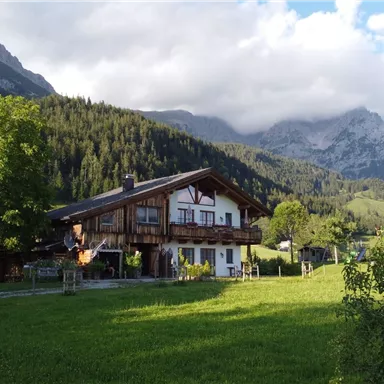 Ein gemütliches Haus in den Bergen, umgeben von grünen Wiesen und majestätischen Gipfeln. Die Szenerie vermittelt einen ruhigen, ländlichen Charme.