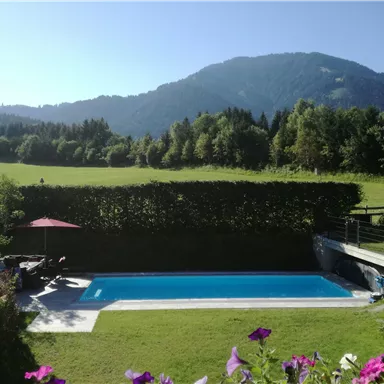 Ein schöner Swimmingpool umgeben von grünen Wiesen und Bäumen. Im Hintergrund sind sanfte Hügel und eine klare blaue Himmel sichtbar.