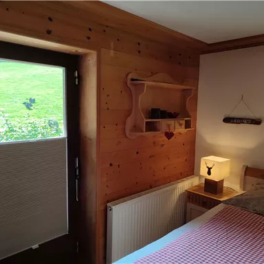 A Cozy room with wooden walls and a large window. On the table stands a lamp and the bedding has a checkered pattern.