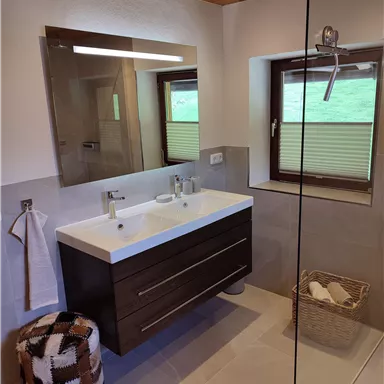 A modern bathroom with a large, bright shower and a two-sink vanity. The warm wooden ceiling and large windows provide natural lighting.