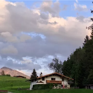 Ein malerisches Haus in der Natur, umgeben von Bäumen und Wiesen. Im Hintergrund sind sanfte Berge und wechselhaft bewölkter Himmel zu sehen.
