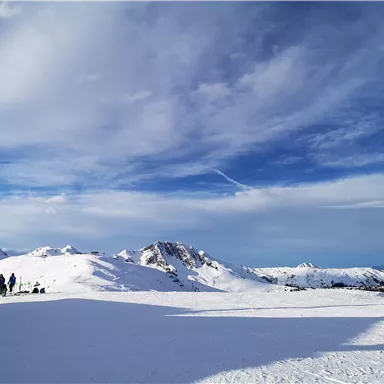 A winter landscape with snow-covered mountains and a clear sky. In the foreground, some people can be seen skiing or hiking.