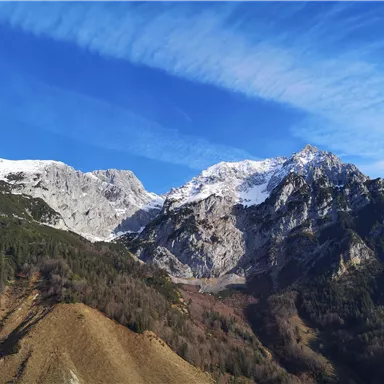 An impressive mountain landscape with snow-covered peaks and a bright blue sky. In the foreground, green hills and forests are visible.