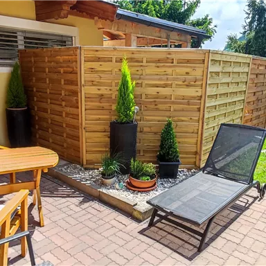 Ein gemütlicher Terrassenbereich mit einem Tisch, Stühlen und einer Liege. Umgeben von grünen Pflanzen und einem Sichtschutz aus Holz.