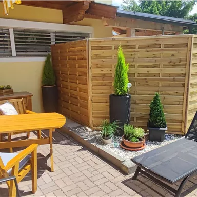 Ein gemütlicher Außenbereich mit einem Tisch, Stühlen und einer Liege. Umgeben von Pflanzen und einer Holzsichtschutzwand.