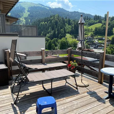 Eine sonnige Terrasse mit einem Liegestuhl, einem kleinen Tisch und einem Sonnenschirm. Im Hintergrund sind grüne Hügel und Wohngebäude sichtbar.