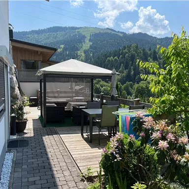 Eine gemütliche Terrasse mit einem Pavillon, umgeben von bunten Blumen und Pflanzen. Im Hintergrund sind grüne Berge und ein blauer Himmel zu sehen.
