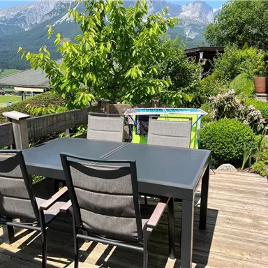 Ein gemütlicher Außenbereich mit einem schwarzen Tisch und vier Stühlen. Im Hintergrund sind schön angelegte Pflanzen und eine beeindruckende Berglandschaft zu sehen.