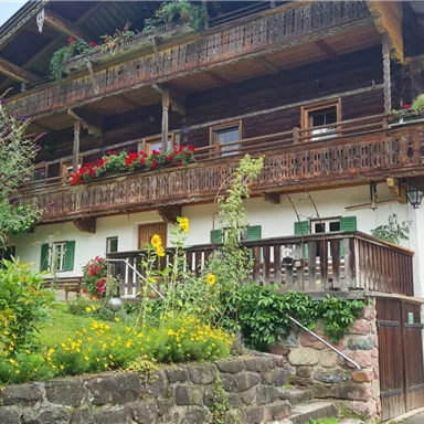 A traditional farmhouse with a wooden porch and blooming flowers. The garden is green and inviting.