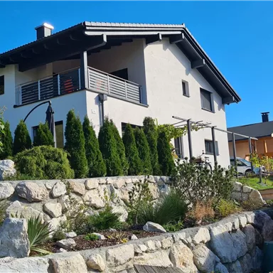 A modern house with a beautiful facade and a garden. The surroundings are green and well-kept, with a stone wall in the foreground.
