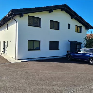 A modern two-story house with a bright facade stands in a quiet environment. A car is parked on the paved driveway in front of the building.