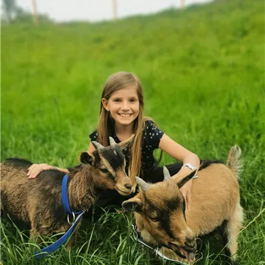 Ein Mädchen sitzt im Gras und spielt mit zwei Ziegen. Die Ziegen sind freundlich und neugierig.