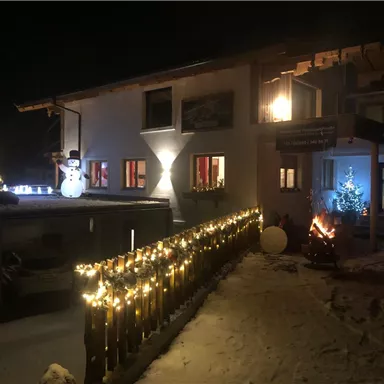 Ein festlich dekoriertes Haus im Schnee, beleuchtet mit Weihnachtslichtern. Auf dem Balkon stehen ein Schneemann und ein geschmückter Weihnachtsbaum.