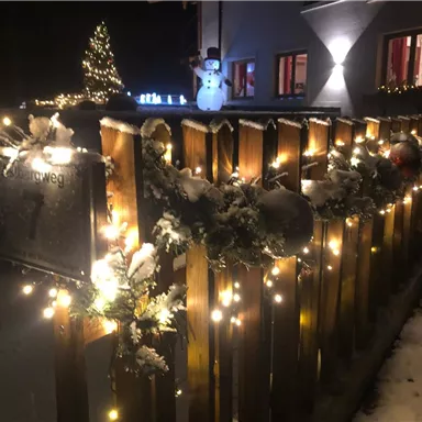Ein festlich geschmückter Zaun mit Lichtern und Weihnachtsdekoration im Schnee. Im Hintergrund steht ein Schneemann und ein Weihnachtsbaum leuchtet.