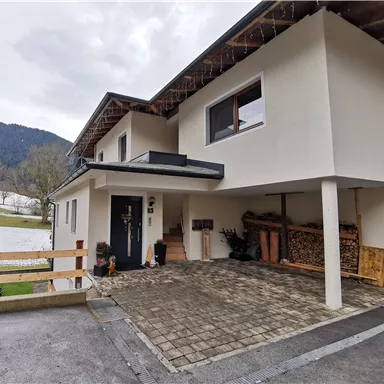 Ein modernes Haus mit einer Einfahrt und einem Parkplatz. Im Hintergrund sind Hügel und ein bewölkter Himmel sichtbar.