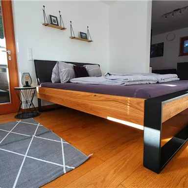 A modern bedroom with a wooden bed and a gray carpet. The room has large windows that let in plenty of light.