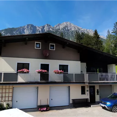 Ein modernes Haus mit einem Balkon, umgeben von grünen Wiesen und Bergen im Hintergrund. Zwei Garagentore und ein blauer Wagen sind ebenfalls sichtbar.