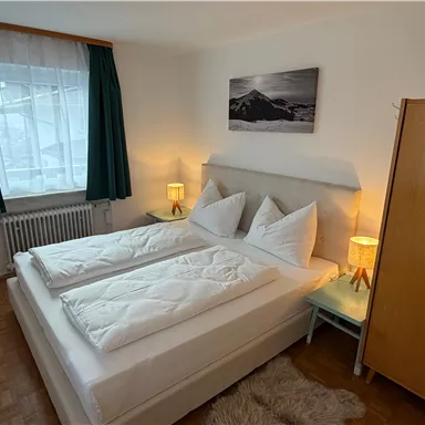 A cozy bedroom with a double bed and two nightstands. The room has large windows and a modern wardrobe.