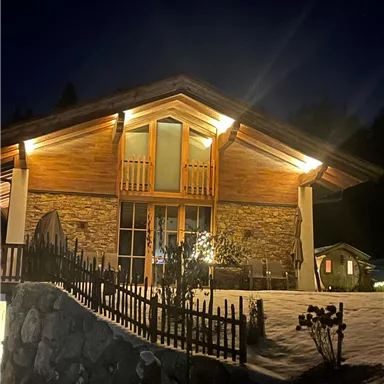 Ein gemütliches Holzhaus in der Nacht, beleuchtet von warmem Licht. Es steht in einer verschneiten Umgebung mit einem kleinen Garten und einem Zaun.