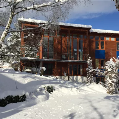 A modern wooden house in the snow, surrounded by a winter landscape. The sky is clear and blue, and the trees are covered with snow.