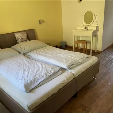 A cozy bedroom with a large bed and two pillows. Next to the bed is a dressing table with a chair.