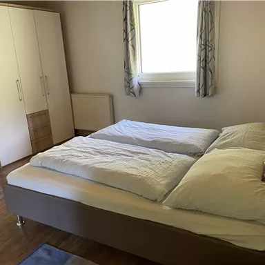 A bright bedroom with a double bed and a large window. The room has a simple style with wooden flooring and a wardrobe.