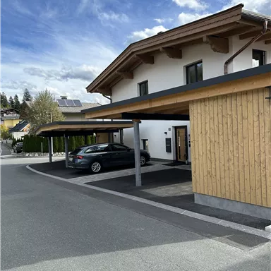 Ein modernes Haus mit Holzverkleidung und einem überdachten Parkplatz. Der Himmel ist blau mit einigen Wolken.