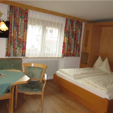 A cozy bedroom with a double bed and a table with chairs. The windows are decorated with colorful curtains and there is a TV on the wall.