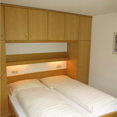 A bright bedroom with a double bed and wooden wardrobes. The bedding is light striped and the lighting is warm and inviting.
