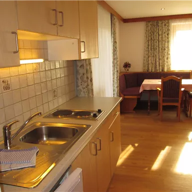 A cozy kitchen with wooden cabinets and a small dining area. Daylight streams through the window, creating a friendly atmosphere.