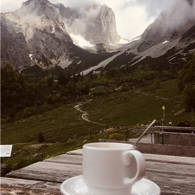 A cup of coffee on a wooden table with a majestic mountain landscape in the background. Fog surrounds the peaks and nature is wonderfully green.
