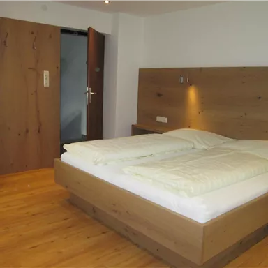 A cozy bedroom with a large double bed and wooden furniture. The floor is made of wood, and a door leads to an adjoining room.