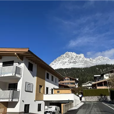 Ein modernes Haus mit Balkonen in einer malerischen Landschaft. Im Hintergrund sind schneebedeckte Berge und ein klarer blauer Himmel zu sehen.