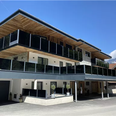 A modern building with glass balconies and wooden details. The facade is bright, surrounded by a picturesque mountain landscape.